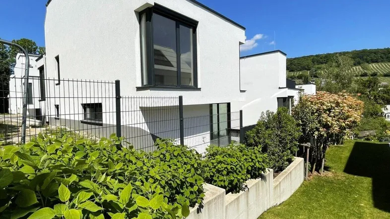 Moderne weiße Architektur mit großen Fenstern, umgeben von grüner Landschaft und Weinbergen unter blauem Himmel.
