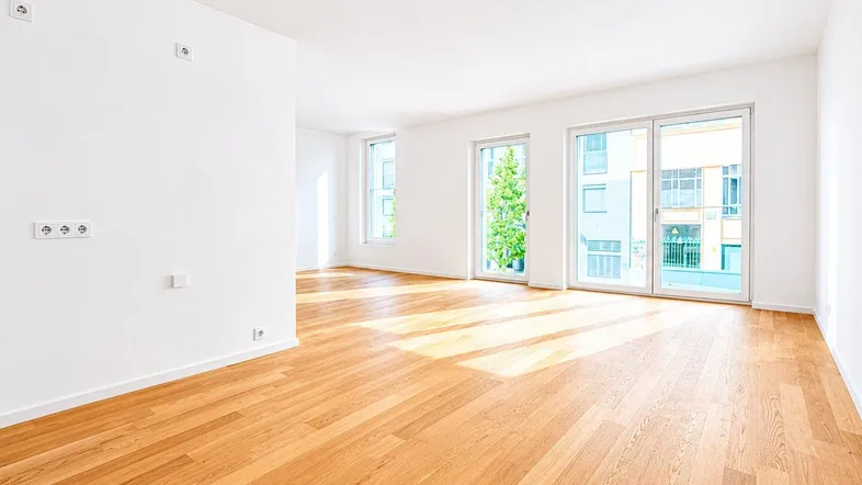 Helles, leeres Zimmer mit Parkettboden und großen Fenstern, die viel Tageslicht hereinlassen.