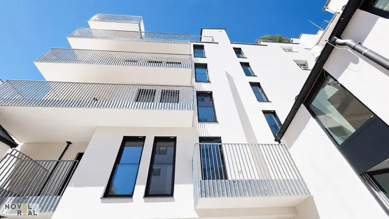 Moderne weiße Fassade eines Neubaus mit mehreren Balkonen und großen Fenstern unter blauem Himmel.