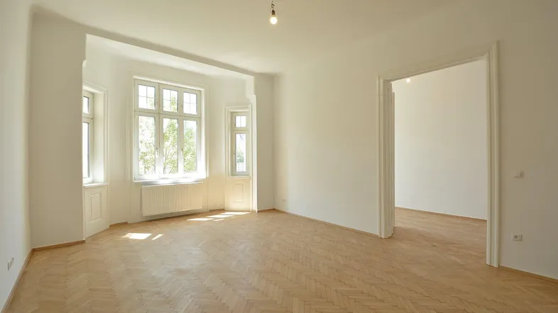 Helles, frisch renoviertes Zimmer mit Fischgrätparkett, hohen Decken und großen Fenstern, die viel Tageslicht hereinlassen.