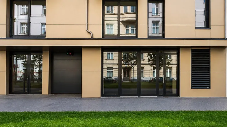 Moderne Gebäudefassade im Erdgeschoss mit großen Fenstern, Garagentor und gepflegter Grünfläche.