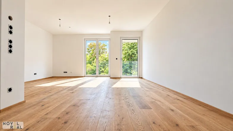 Heller, leerer Wohnraum mit Holzboden und großen Fenstern, die viel Tageslicht hereinlassen.