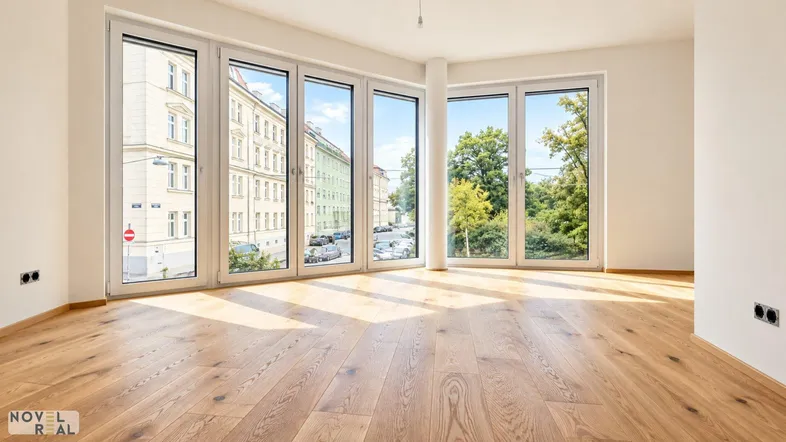 Helles, leeres Zimmer mit großen Fenstern, Holzboden und Blick auf die Straße und Bäume.