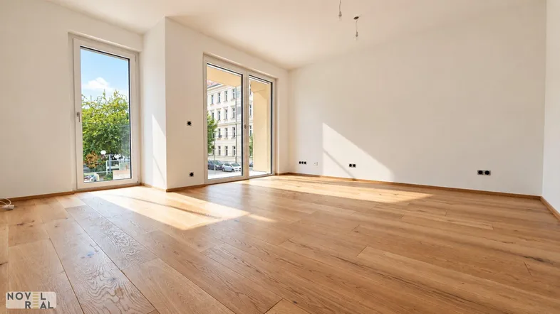 Helles, leeres Zimmer mit Holzboden und großen Fenstern, die viel Tageslicht hereinlassen.
