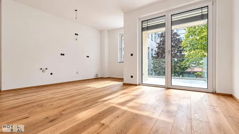 Heller, leerer Raum mit Holzboden und großen Fenstern, die Zugang zu einem Balkon bieten.