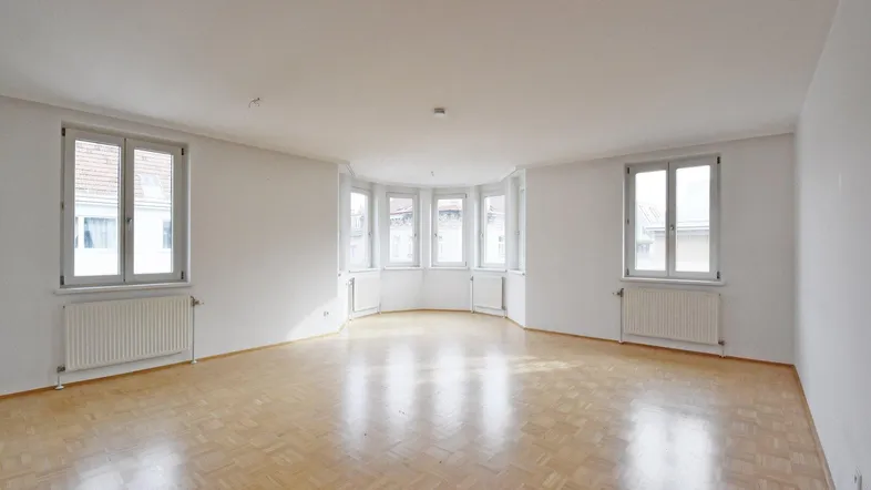 Helles Wohnzimmer mit Parkettboden und einem Erkerfenster, das viel Tageslicht hereinlässt.