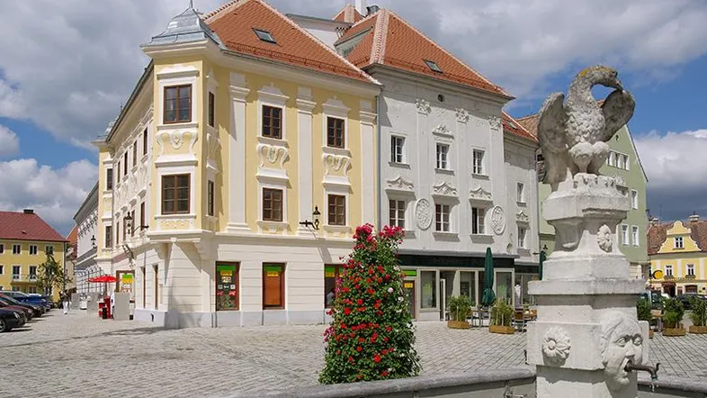 Historisches Gebäude an einem belebten Stadtplatz mit einem dekorativen Brunnen und Blumenarrangement.
