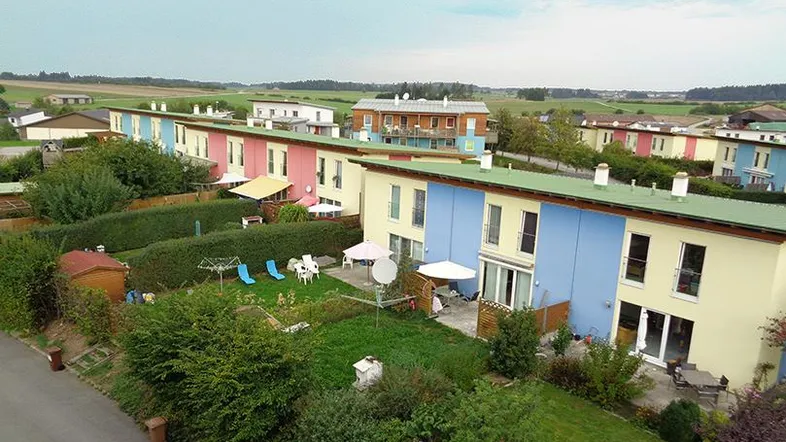 Luftaufnahme einer modernen Reihenhaussiedlung mit farbenfrohen Fassaden und privaten Gärten in ländlicher Umgebung.