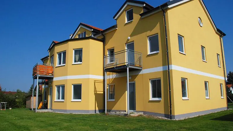 Gelbes Mehrfamilienhaus mit mehreren Balkonen und grüner Rasenfläche unter blauem Himmel.