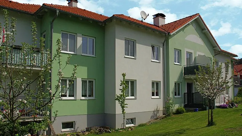 Mehrfamilienhaus mit hellgrüner und beiger Fassade, roten Ziegeldächern und gepflegtem Vorgarten.