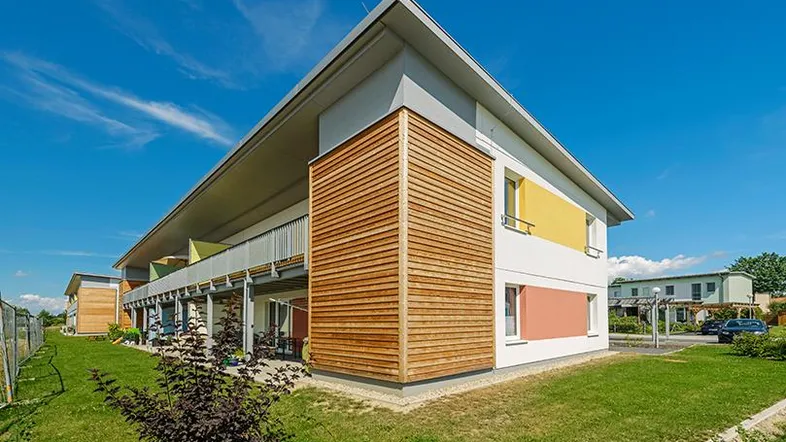 Modernes Mehrfamilienhaus mit farbigen Akzenten, Holzverkleidung und gepflegter Grünfläche unter blauem Himmel.