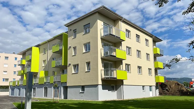 Modernes Mehrfamilienhaus mit hellgelber Fassade, leuchtend grünen Balkonen und gepflegter Grünfläche unter blauem Himmel.