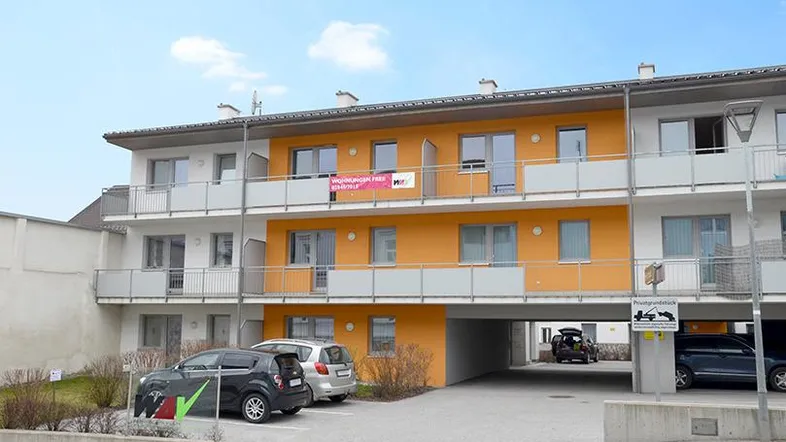 Mehrfamilienhaus mit gelber Fassade, Balkonen und Parkplätzen im Freien unter blauem Himmel.