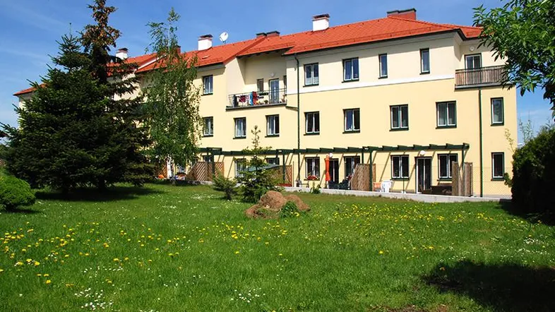 Mehrfamilienhaus mit gelber Fassade, rotem Dach und großem, gepflegtem Rasenbereich.