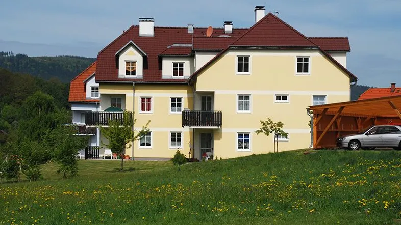Mehrfamilienhaus mit gelber Fassade, rotem Dach und Balkonen in ländlicher Umgebung.