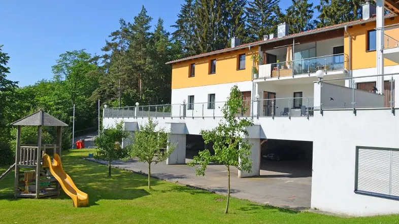 Modernes Mehrfamilienhaus mit Spielplatz, Tiefgaragenzufahrt und Balkonen, umgeben von viel Grün.