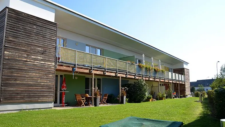 Moderne Wohnanlage mit Holzfassade, Balkonen und einer grünen Rasenfläche mit Sandkasten.