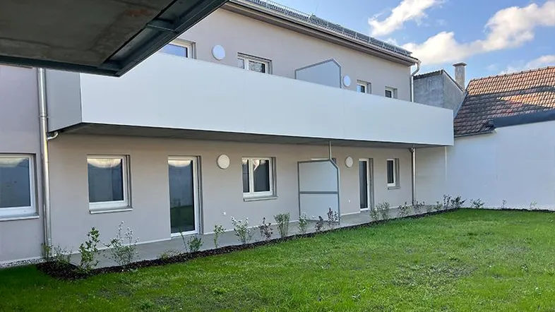 Moderne Neubau-Fassade mit Balkon und kleiner Grünfläche im Vordergrund.