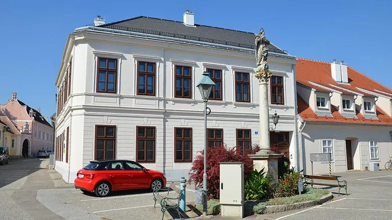 Historisches weißes Gebäude mit braunen Fenstern an einem sonnigen Platz mit einem roten Auto.