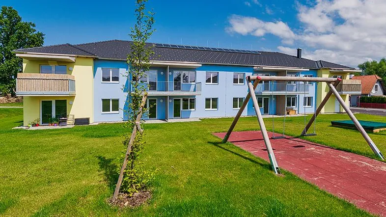 Modernes Mehrfamilienhaus mit farbenfroher Fassade, Balkonen und einem Kinderspielplatz im Vordergrund.