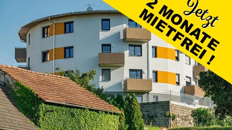 Modernes rundes Apartmentgebäude mit Balkonen und orangefarbenen Akzenten, umgeben von Grünflächen und einem älteren Haus mit Efeubewuchs.