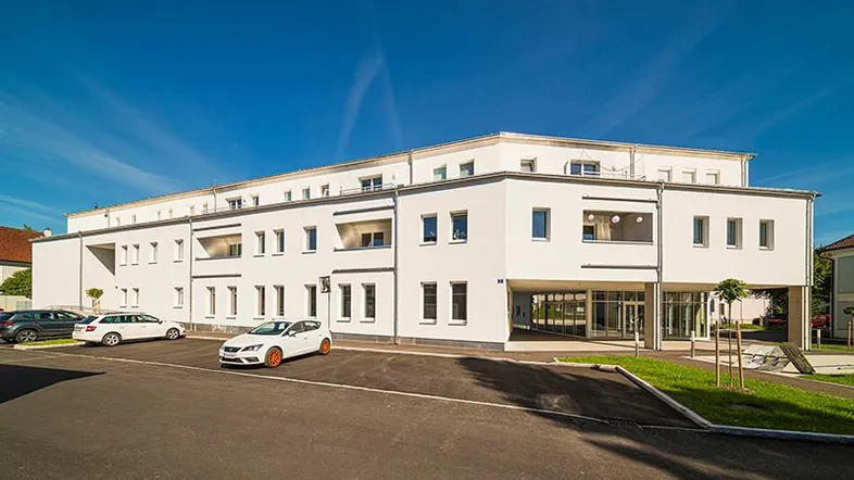 Modernes Mehrfamilienhaus mit weißer Fassade, großen Fenstern und ausreichend Parkplätzen im Freien.