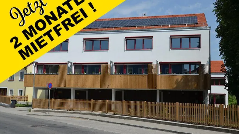Modernes Mehrfamilienhaus mit weißer Fassade, roten Fenstern und Solaranlage auf dem Dach.