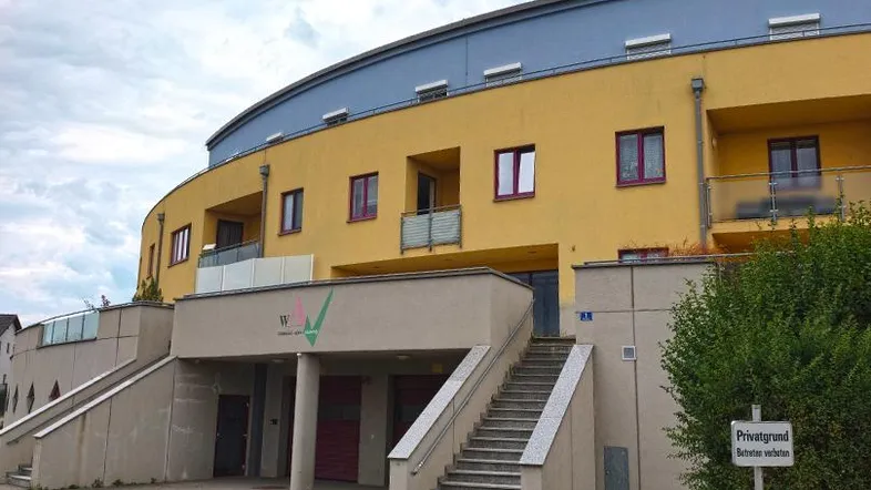 Modernes, geschwungenes Gebäude mit gelber Fassade, blauer Dachpartie und Balkonen.