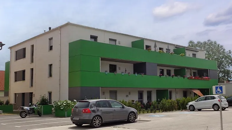Modernes Mehrfamilienhaus mit grüner Fassadengestaltung und barrierefreien Parkplätzen.