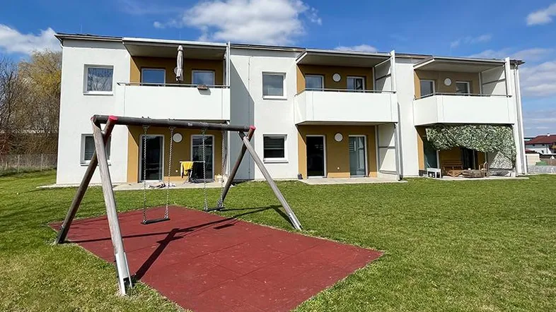 Modernes Mehrfamilienhaus mit Balkonen, umgeben von einer Grünfläche und einem Kinderspielplatz.