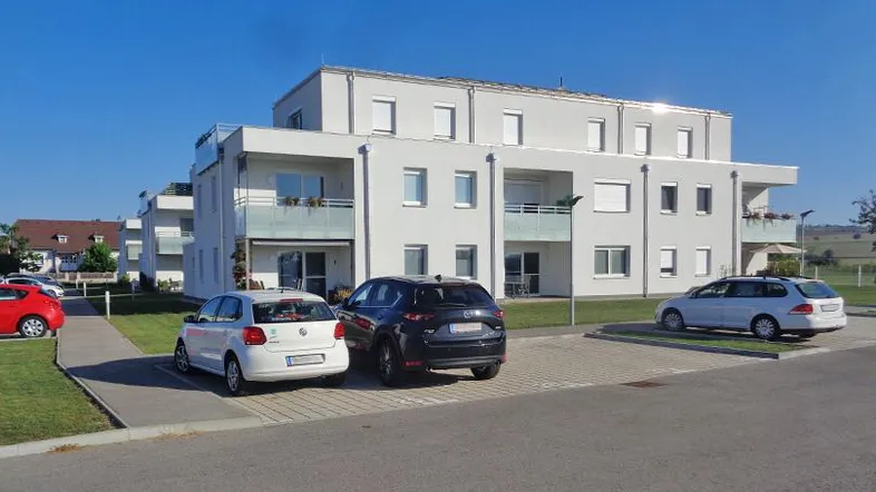Modernes Mehrfamilienhaus mit weißer Fassade, Balkonen und Außenstellplätzen in ländlicher Umgebung.