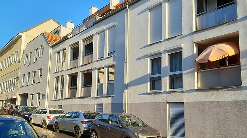Ansicht eines modernen Wohngebäudes mit weißer Fassade, Rollläden und einem kleinen Balkon an einer städtischen Straße.