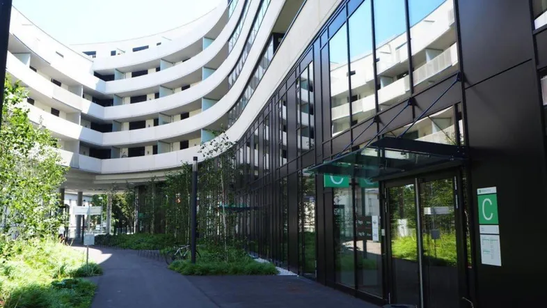 Modernes Gebäude mit geschwungener weißer Fassade, großen Glasflächen und begrüntem Außenbereich.