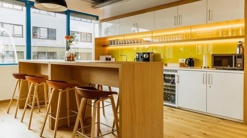 Moderne Büroküche mit Holzboden, weißen Hochglanzschränken und gelber Rückwand, ideal für Pausen.
