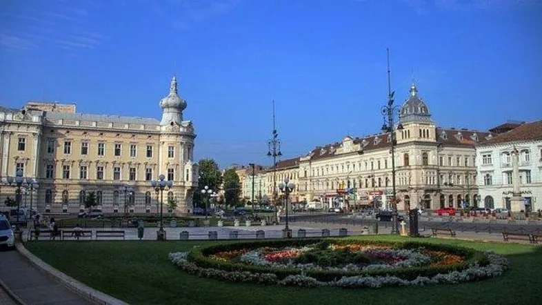 Historischer Stadtplatz mit prächtigen Gebäuden, einem Blumenbeet und blauem Himmel.