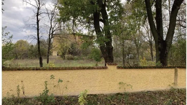 Eine gelbe Mauer mit Ziegelabdeckung trennt eine Wiese von einem bewaldeten Bereich unter bewölktem Himmel.