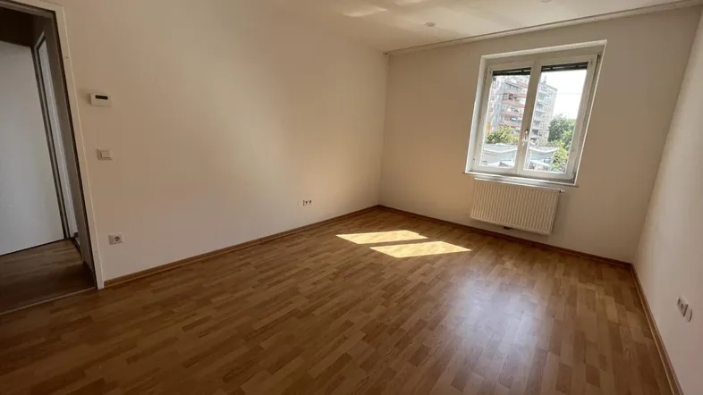 Helles, leeres Zimmer mit Parkettboden und einem Fenster, das Tageslicht hereinlässt.