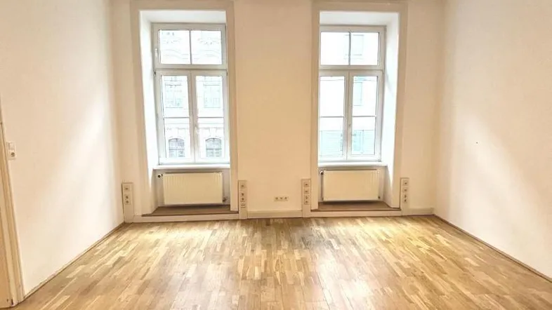 Heller, leerer Raum mit Holzboden und hohen Decken, beleuchtet durch zwei große Fenster.
