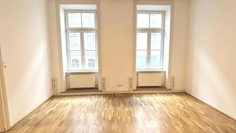 Helles Zimmer mit Parkettboden und zwei großen Fenstern, ideal für Wohn- oder Arbeitszwecke.
