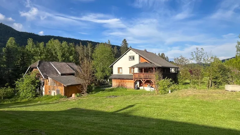 Zwei traditionelle Häuser in ländlicher Umgebung mit grünen Wiesen und bewaldeten Hügeln unter blauem Himmel.