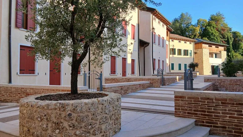 Mediterraner Innenhof mit Olivenbaum, Steinmauern und traditionellen Gebäuden mit roten Fensterläden.
