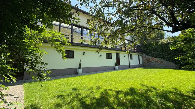 Hausfassade mit großem Rasenbereich und einem verglasten Wintergarten im Obergeschoss, umgeben von Bäumen.