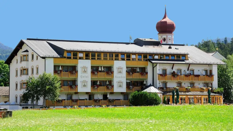 Hotelgebäude mit traditioneller Architektur, Balkonen und einem Turm, umgeben von grünen Wiesen und Bergen.