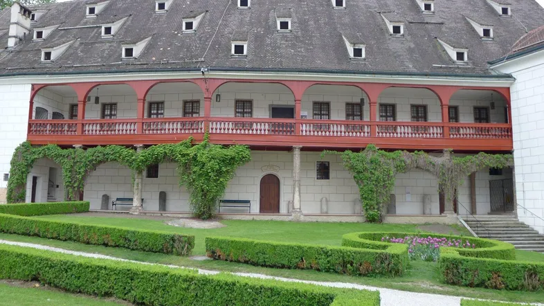 Historisches Gebäude mit rotem Holzbalkon und gepflegtem Gartenbereich mit Formschnitthecken.