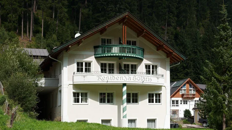 Traditionelles Gebäude mit Balkon und Bergblick, umgeben von grünen Bäumen und Natur.