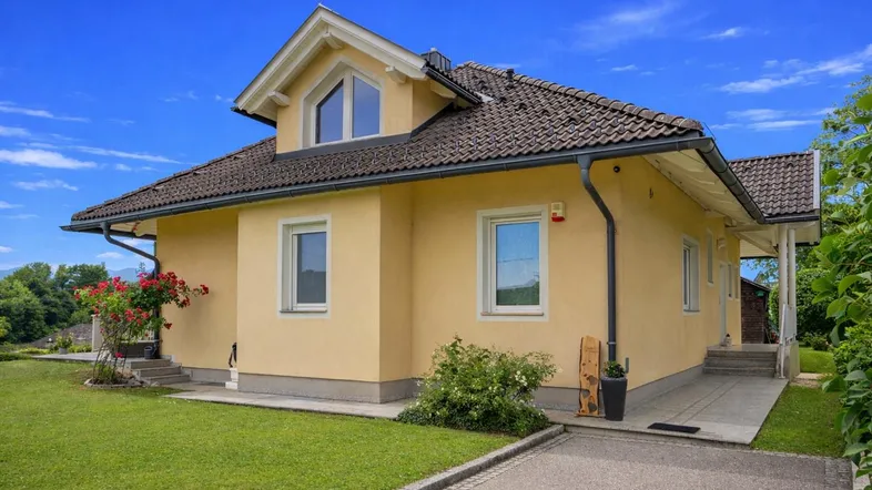 Gelbes Einfamilienhaus mit Satteldach, gepflegtem Vorgarten und blühenden Pflanzen unter blauem Himmel.