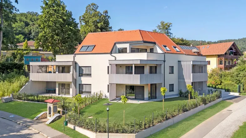 Modernes Mehrfamilienhaus mit rotem Ziegeldach, Balkonen und gepflegter Grünanlage in ländlicher Umgebung.