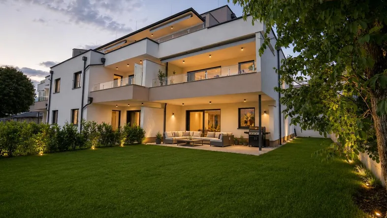 Moderne Wohnanlage bei Dämmerung mit beleuchteter Terrasse, Garten und Balkonen in den oberen Etagen.