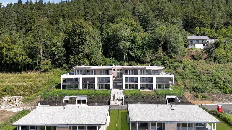 Moderne Wohnanlage mit mehreren Gebäuden, eingebettet in eine grüne Hügellandschaft mit Wald.