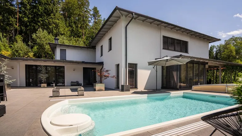 Modernes Haus mit großem Pool und weitläufiger Terrasse, umgeben von grünen Bäumen unter blauem Himmel.
